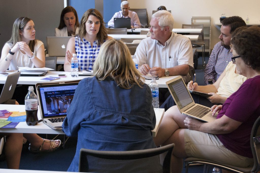 A group of professors sit in a circle listening to a woman talk.